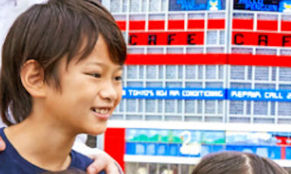 A family of four smiles and points while standing outdoors near a miniature city display on a sunny day.