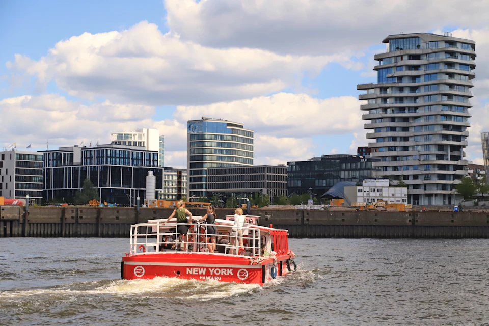 A small red boat with "New York Hamburg" on its stern travels on water with modern buildings and a partly cloudy sky in the background.