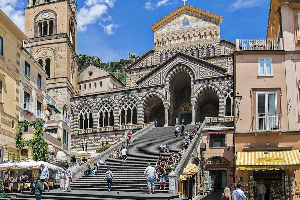 A historic cathedral with ornate architecture and a staircase leading to it, with people sitting and walking in the area.