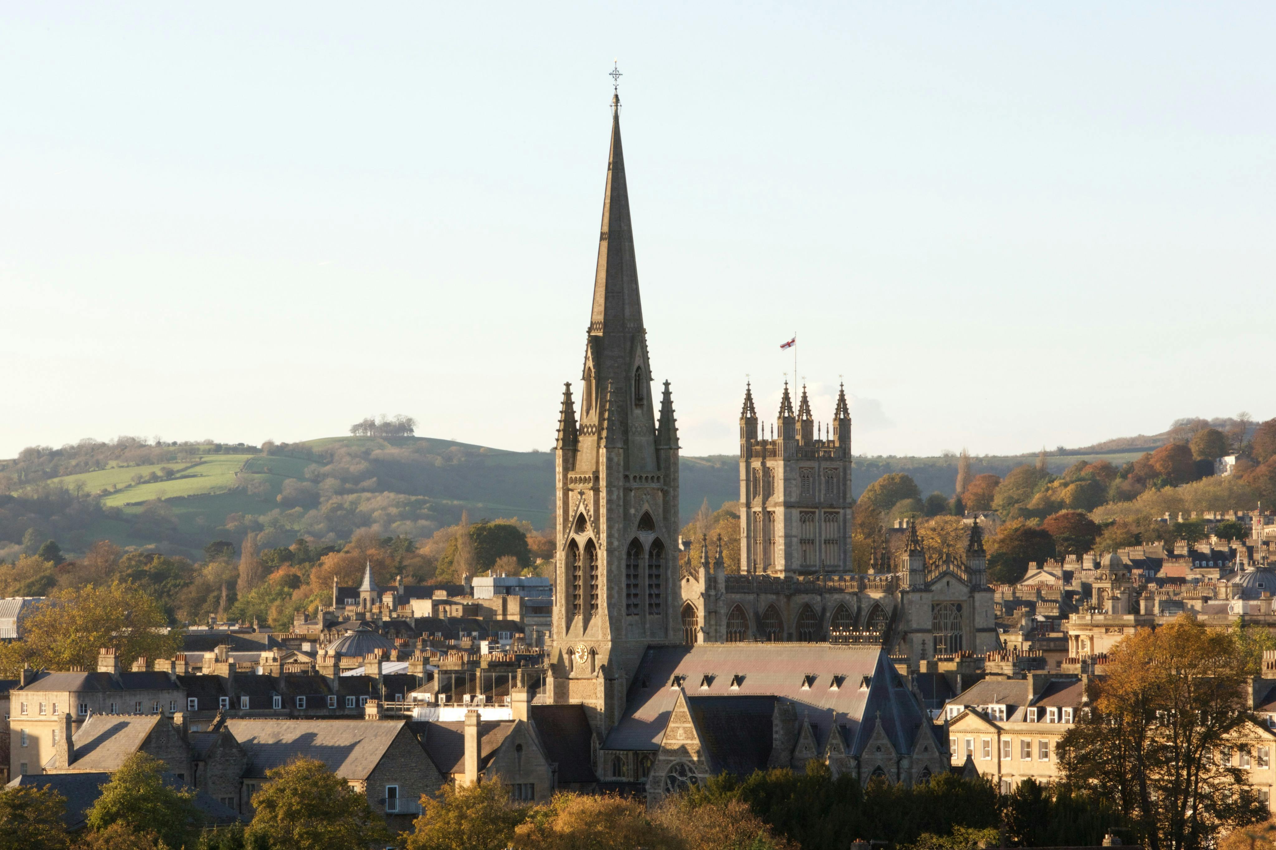 A scenic view of a city with prominent church spires and rooftops, surrounded by trees and hills under a clear sky.