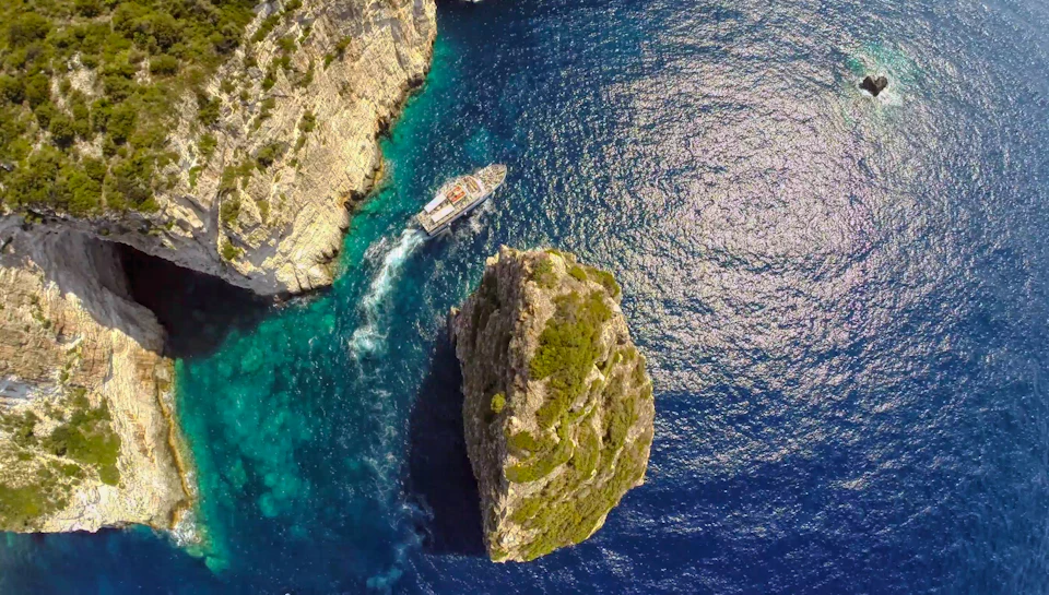 Aerial view of a boat navigating between cliff-lined shore and a small, rocky island in clear blue water.