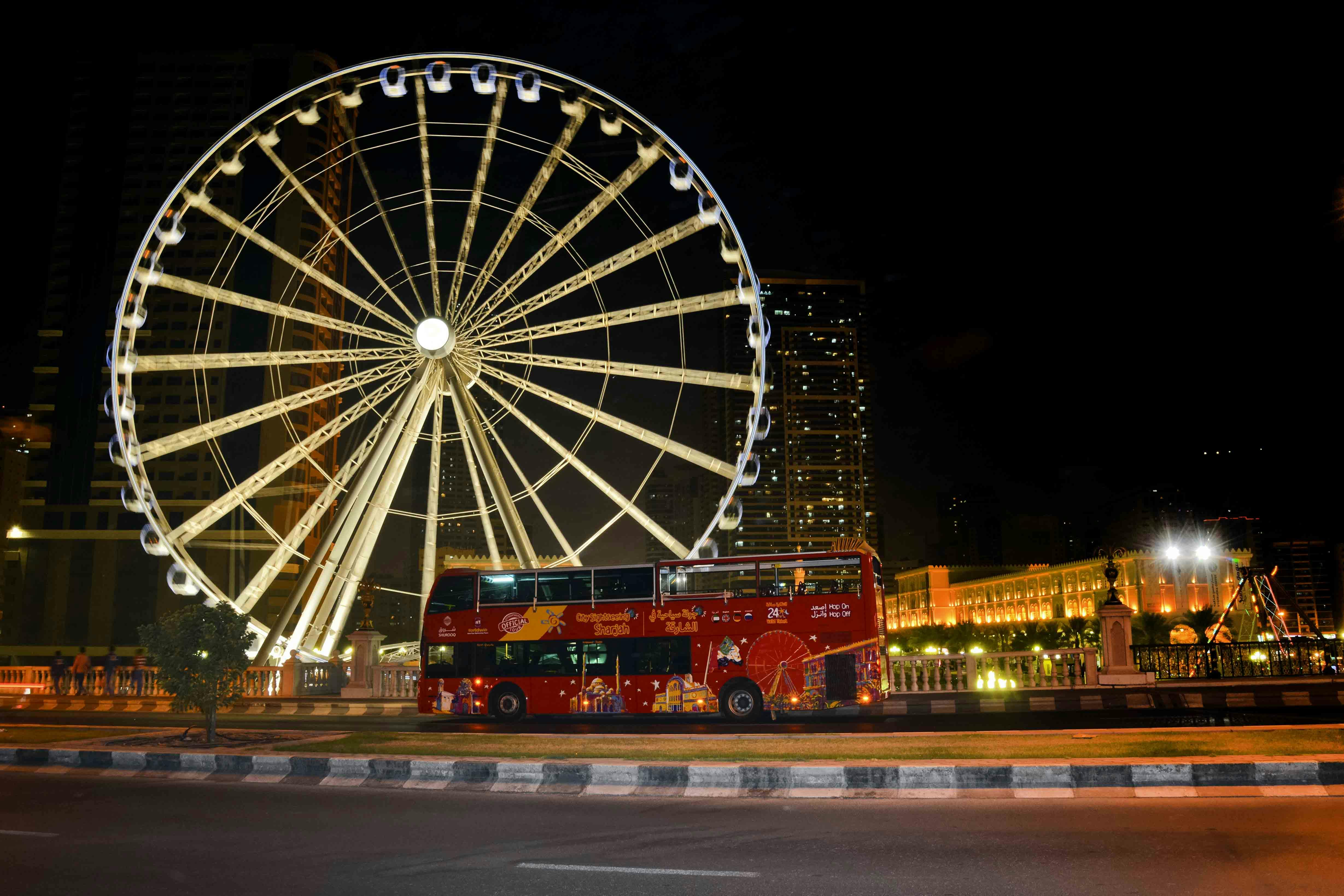A red double-decker tour bus is passing by a large illuminated Ferris wheel at night, with city buildings in the background.