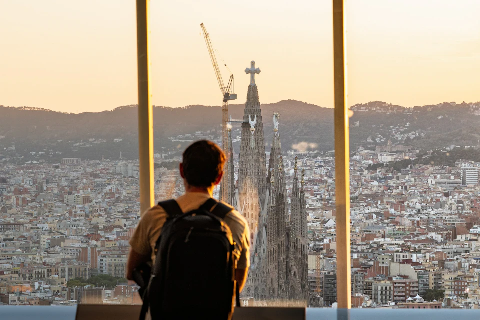 A person with a backpack views a cityscape with a prominent cathedral and crane at sunset through a large window.