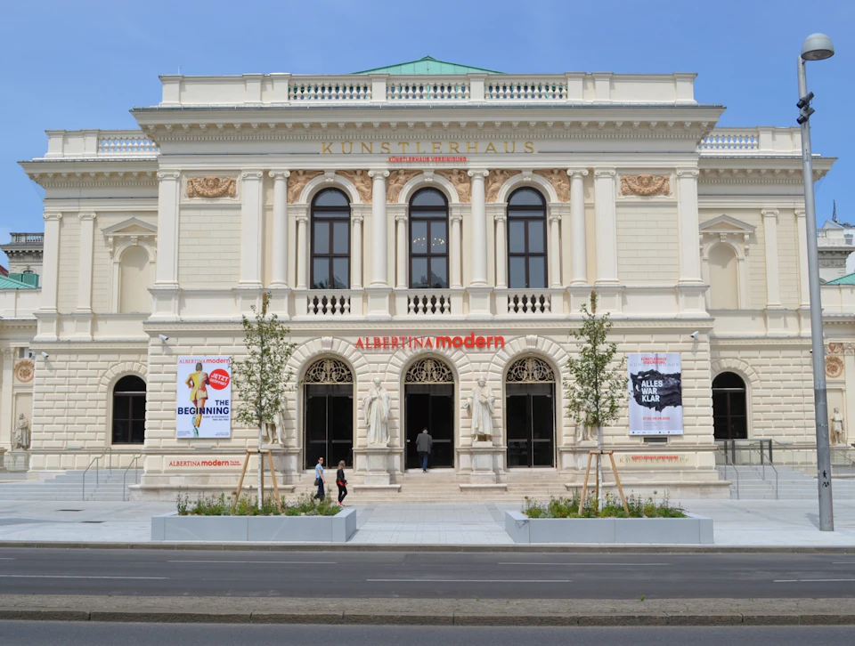 A neoclassical building with three arched entrances, posters on the facade, and trees in planters along the sidewalk.