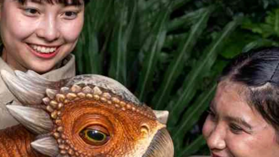 Two people in uniforms interact with a realistic baby dinosaur model in a lush jungle-like setting.
