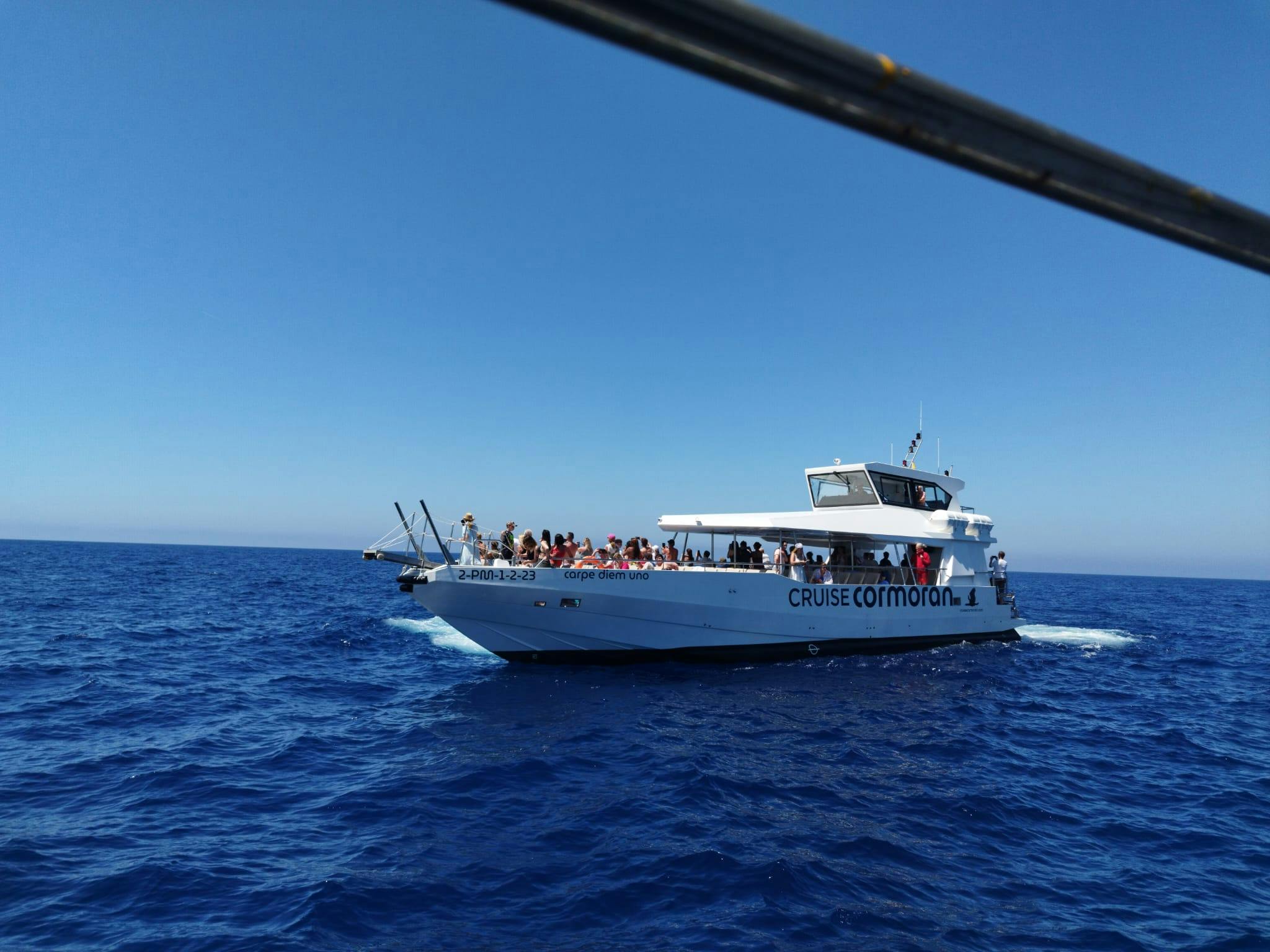 Ένα λευκό κρουαζιερόπλοιο με το όνομα «Cormoran» στην ανοιχτή θάλασσα, γεμάτο επιβάτες κάτω από τον ηλιόλουστο γαλάζιο ουρανό.