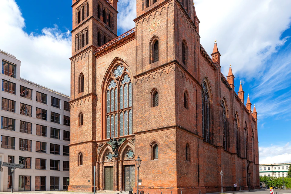 A red brick Gothic-style church with twin towers, pointed arches, and ornate windows under a blue sky with clouds.