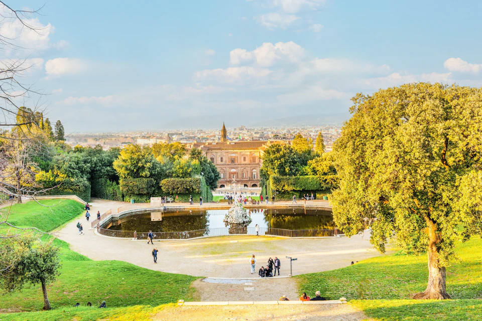A scenic view of a park with a fountain, trees, and a historical building in the background under a blue sky.