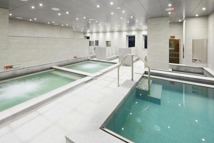 An indoor spa facility with multiple rectangular pools, stainless steel railings, and bright ceiling lights.