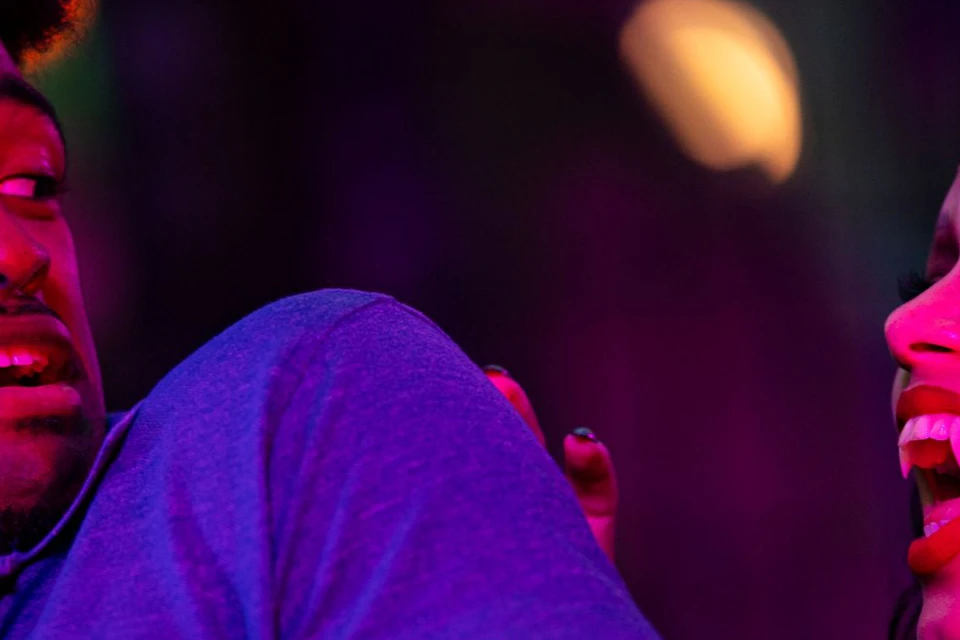 A man in a blue shirt reacts with surprise as a woman in makeup and jewelry gestures animatedly under purple lighting.