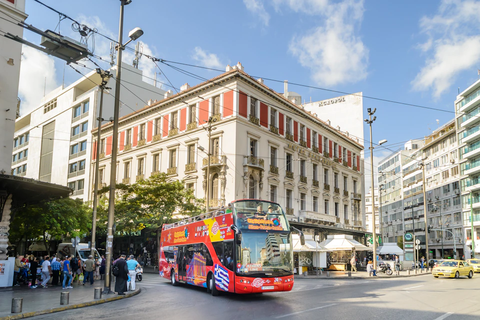 A red double-decker tour bus is seen on a city street, passing by a large, ornate building with red and white accents.