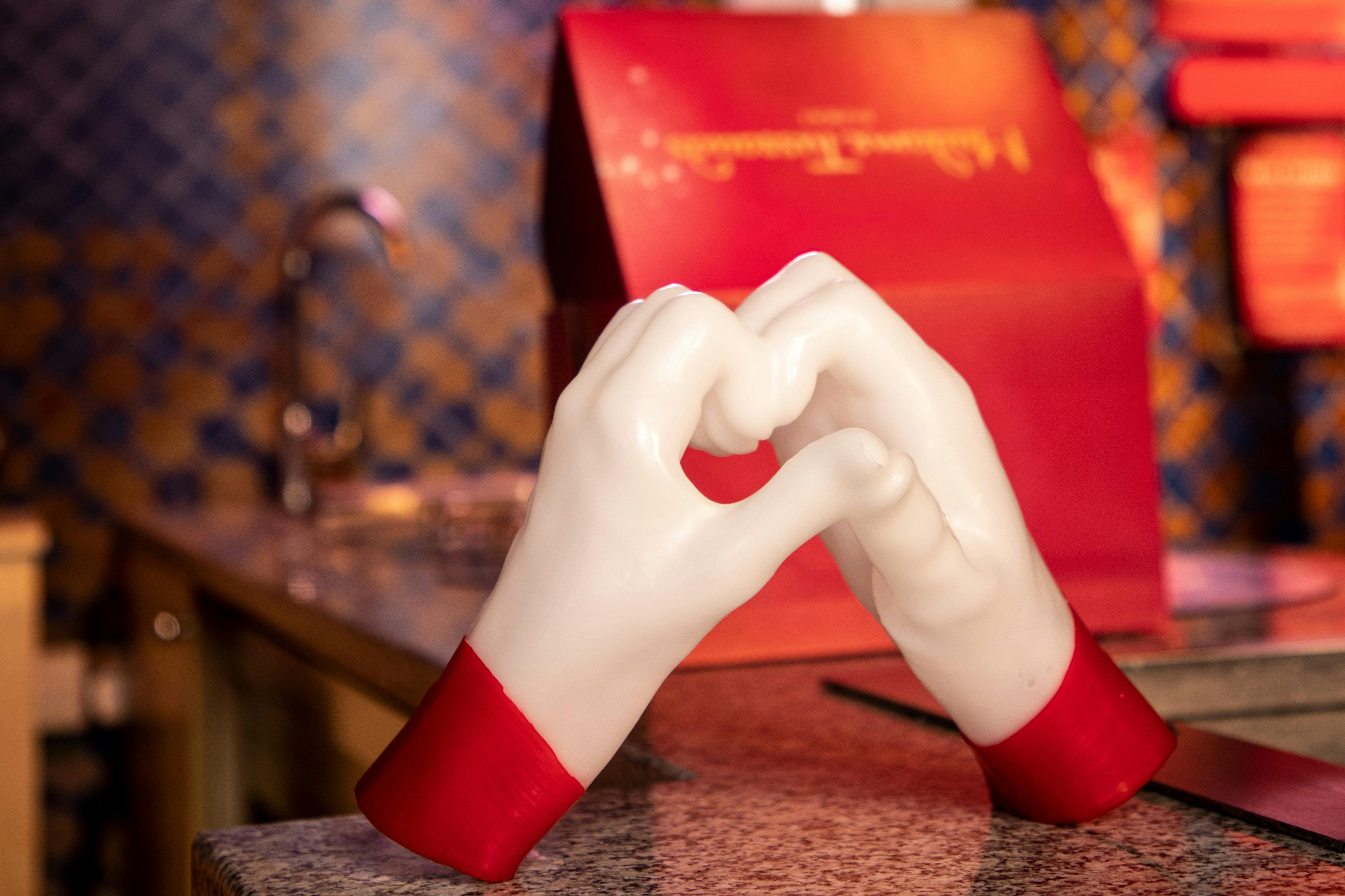 Δύο χέρια από λευκό κερί σχηματίζουν ένα σχήμα καρδιάς σε έναν πάγκο με ένα θολό κόκκινο κουτί και καρό φόντο.