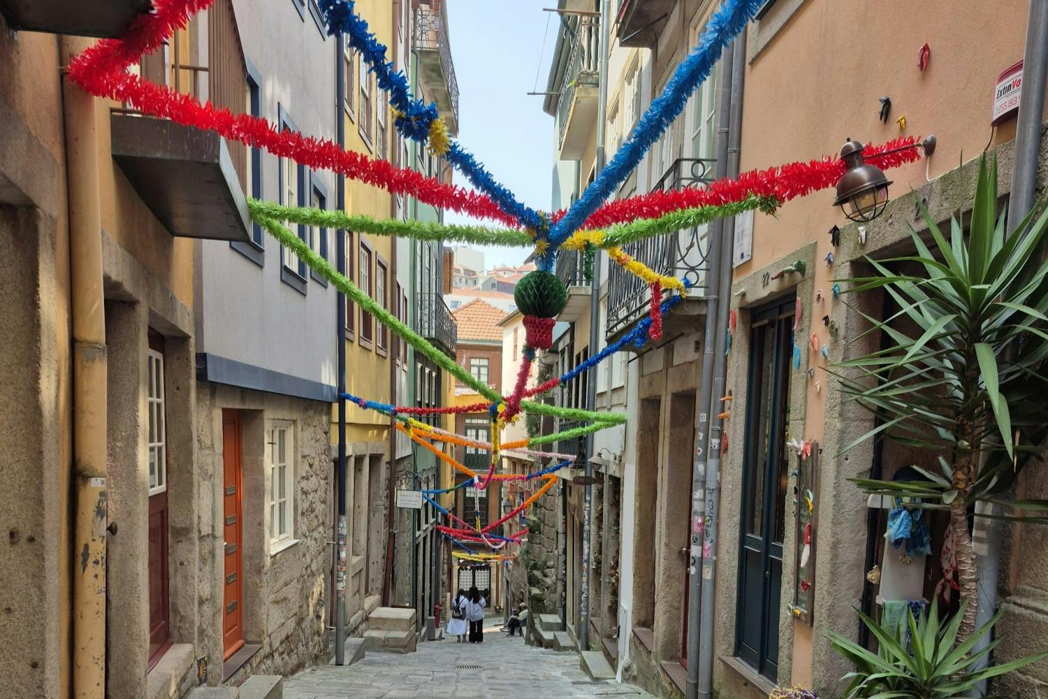 Eine schmale, farbenfrohe Gasse mit festlichen Girlanden über dem Kopf, gesäumt von Häusern und zwei Menschen, die unten durchgehen.