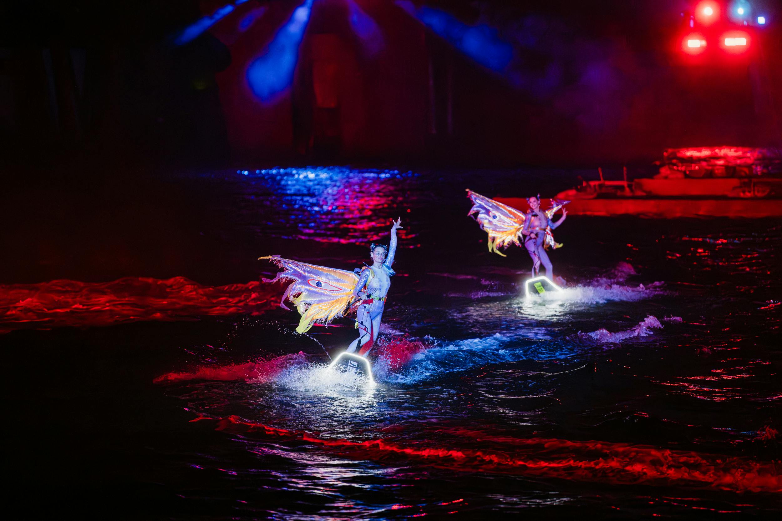 Two performers on illuminated water jetboards, wearing colorful costumes, in a nighttime show with dramatic lighting effects.