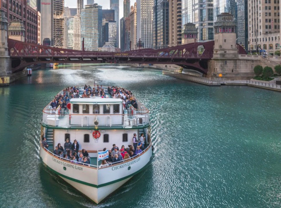 A riverboat named "Chicago's Leading Lady" with passengers aboard travels under a bridge in a city with tall buildings.