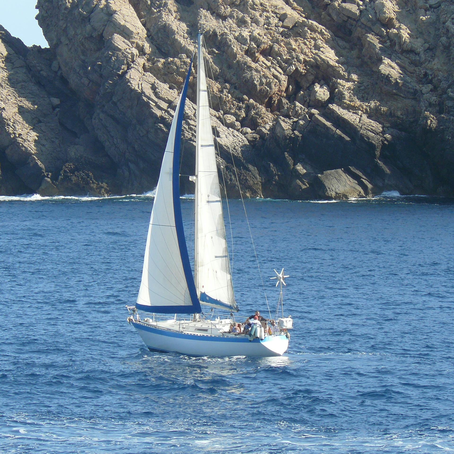 Un voilier aux voiles blanches avec plusieurs personnes à bord naviguant près de falaises rocheuses dans une mer bleue et calme.