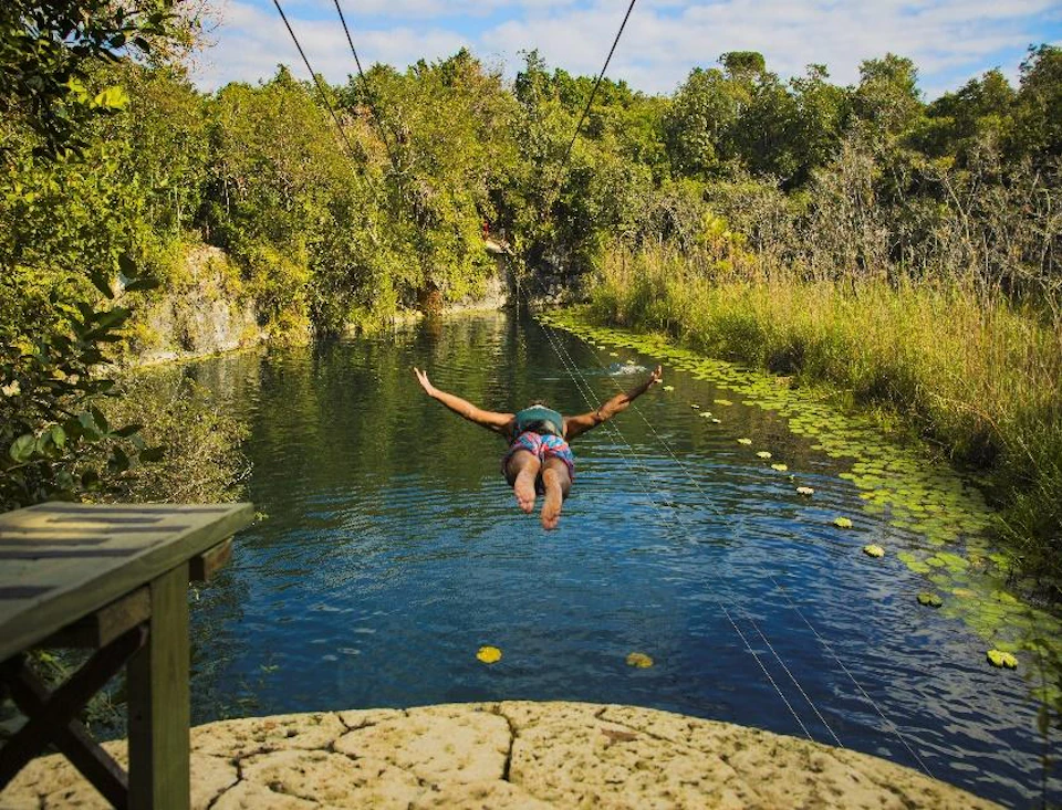 Person ziplining over a clear blue river surrounded by lush green trees and vegetation under a partly cloudy sky.