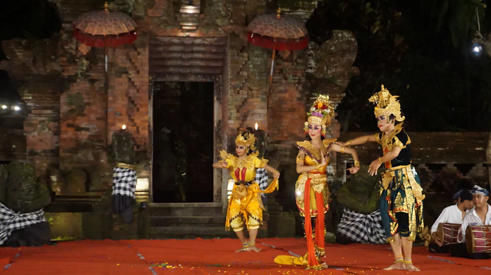 Traditional dancers in ornate costumes perform on a stage with musicians and a temple-like backdrop in the evening.