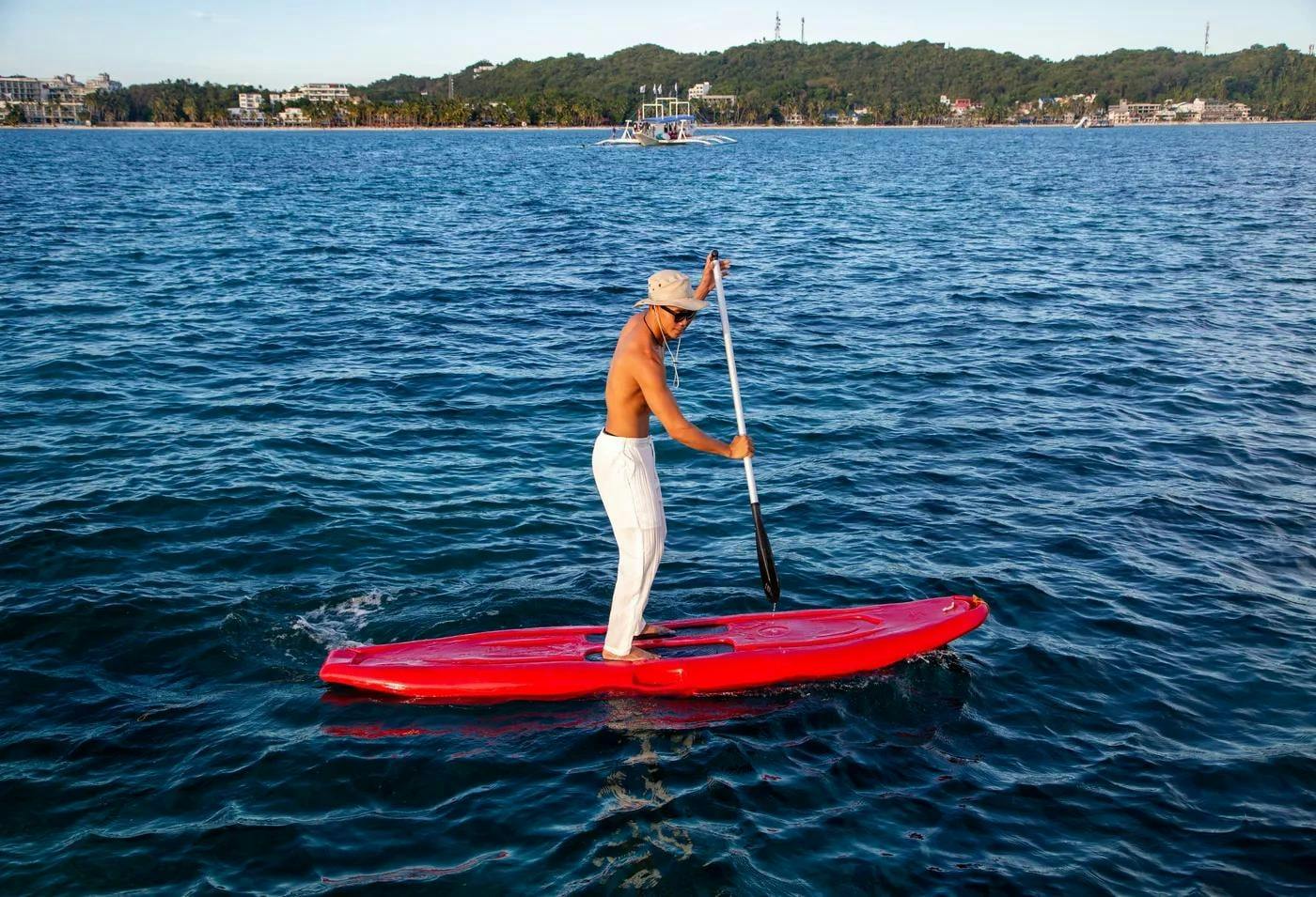 A person paddleboarding on a red board in calm blue water, with a forested shoreline and boats in the background.