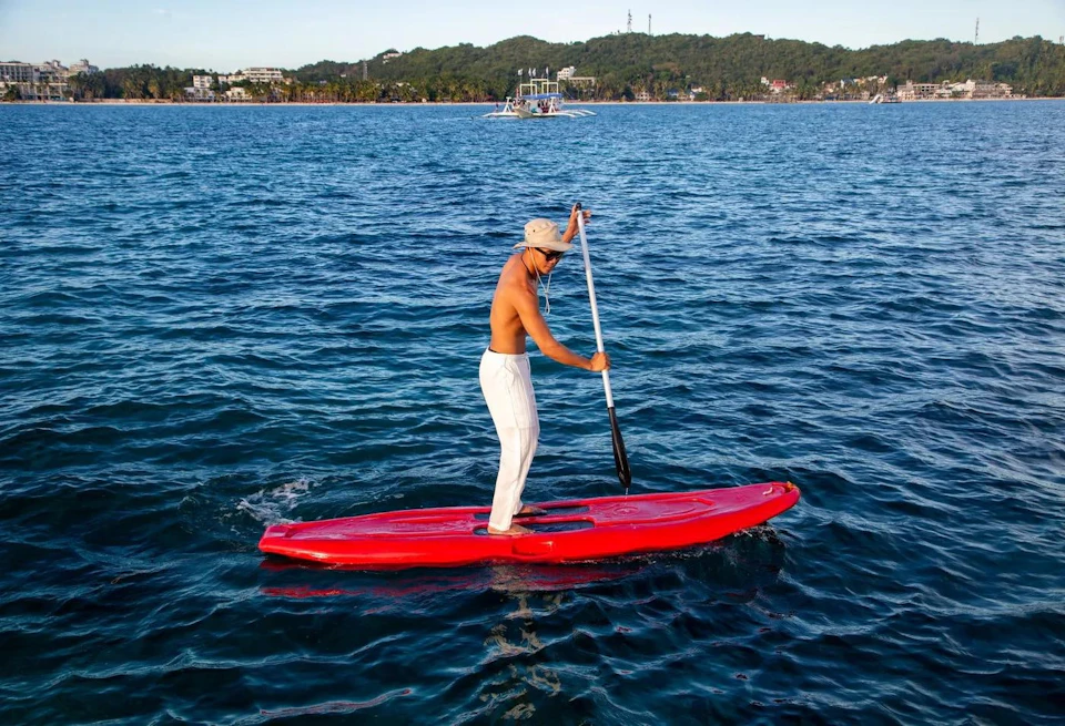 A person paddleboarding on a red board in calm blue water, with a forested shoreline and boats in the background.