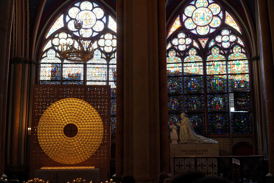 Intricately designed stained glass windows alongside a golden circular altar and statue inside a dimly lit cathedral.