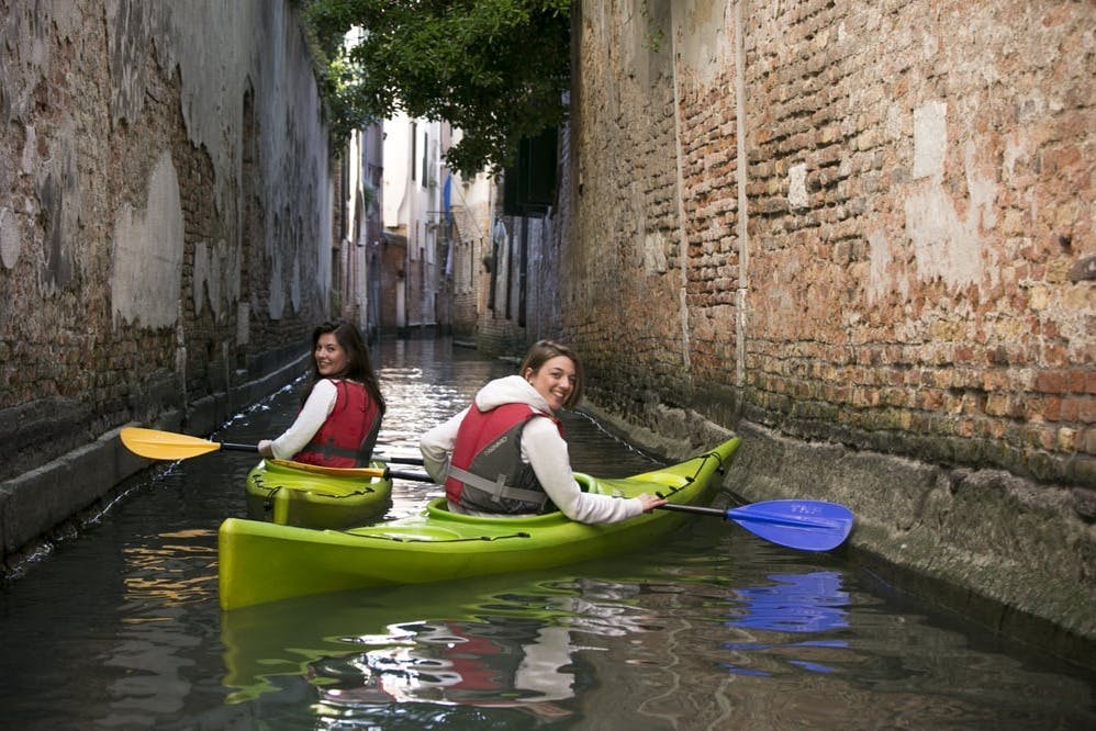 Deux femmes font du kayak dans un canal étroit bordé de murs en briques, en souriant à l'appareil photo.