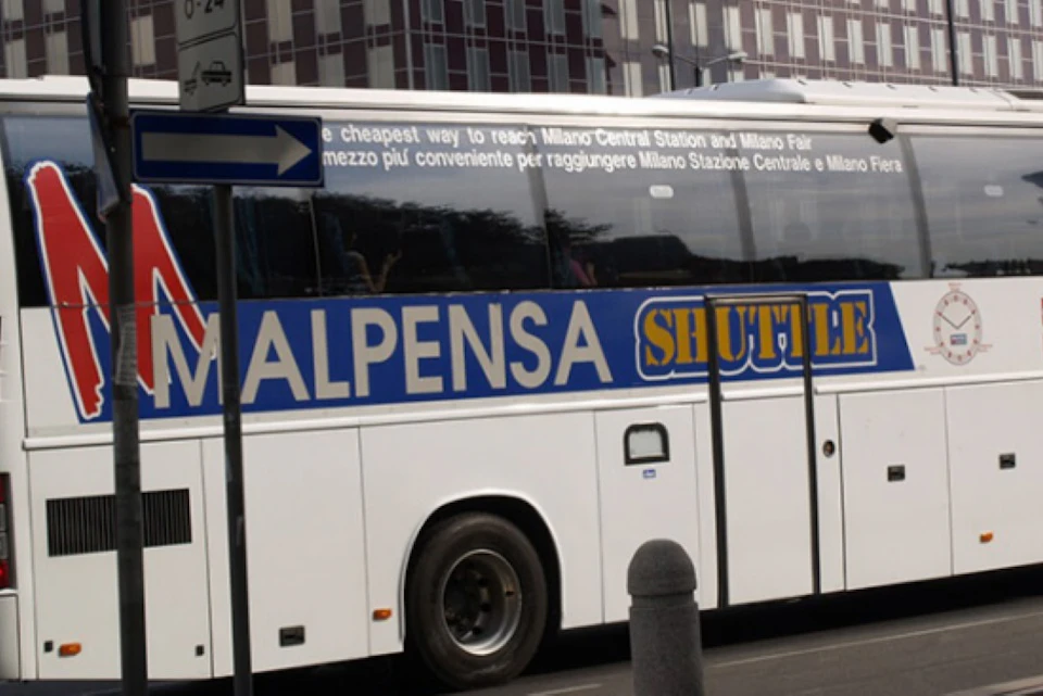 White bus labeled "MALPENSA SHUTTLE" parked on a city street, with buildings and a sign in the background.