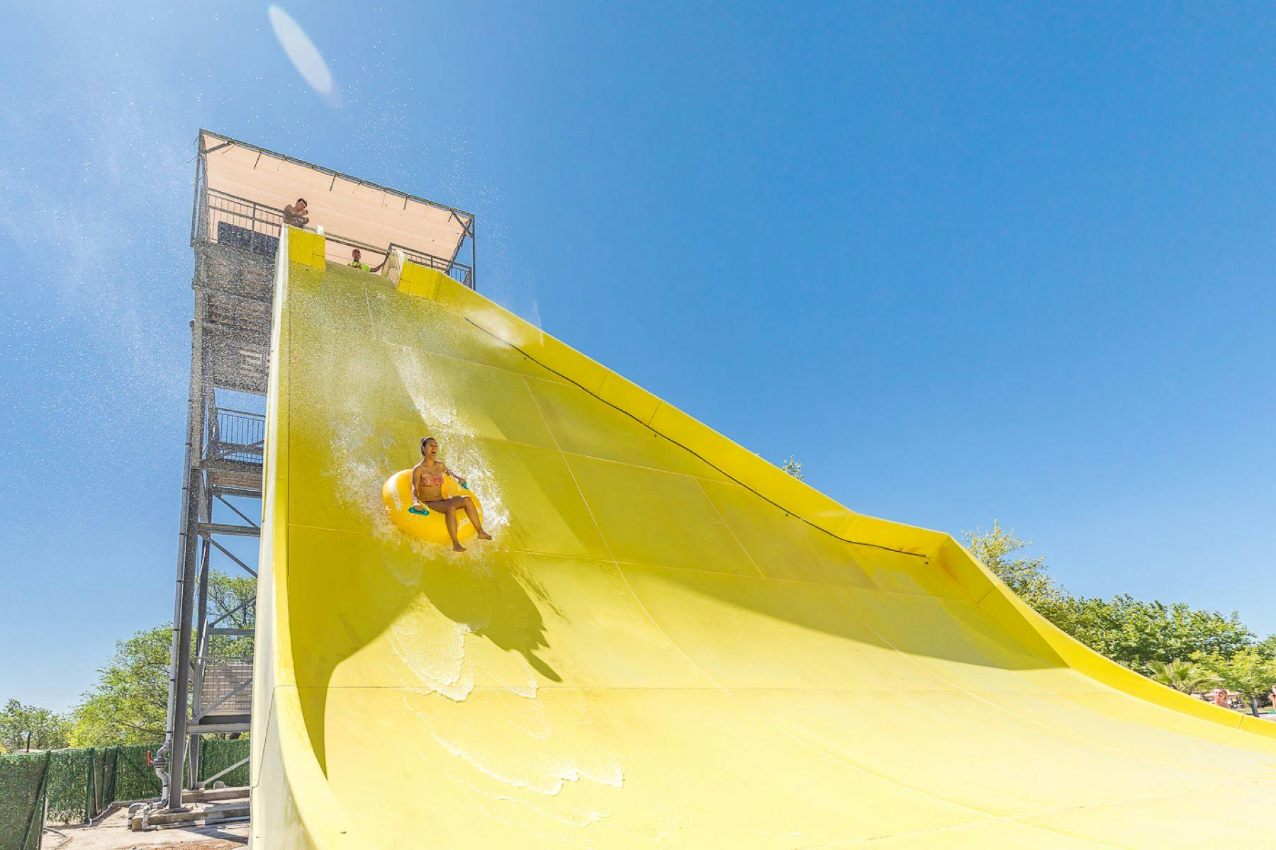 Une personne qui dévale un toboggan aquatique jaune vif sur une bouée gonflable, sous un ciel bleu limpide.