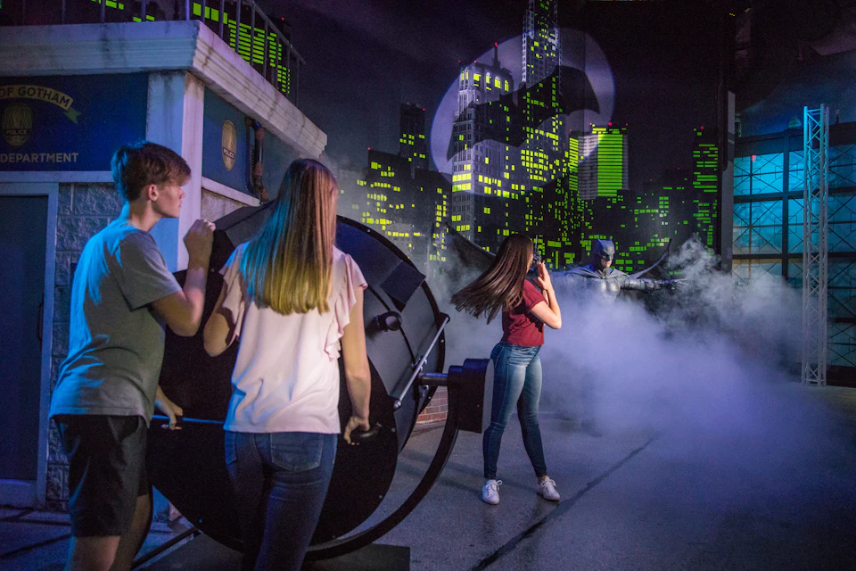 Three people operate a large spotlight with the Batman symbol projected onto a cityscape backdrop.