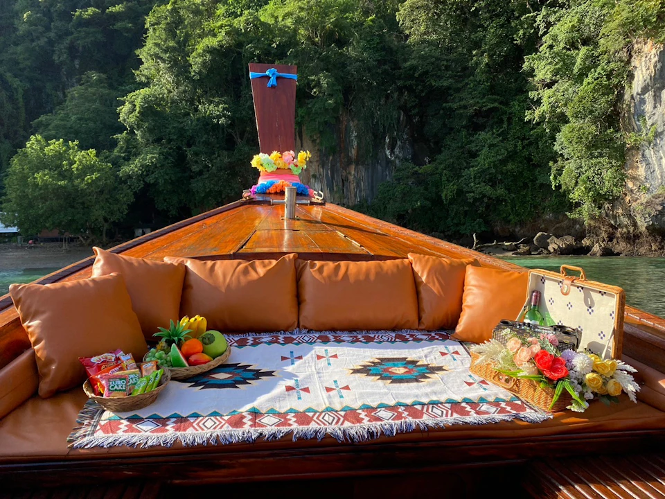 Fruit and snacks on a decorated blanket on a boat's deck, surrounded by brown cushions, with lush green trees in the background.