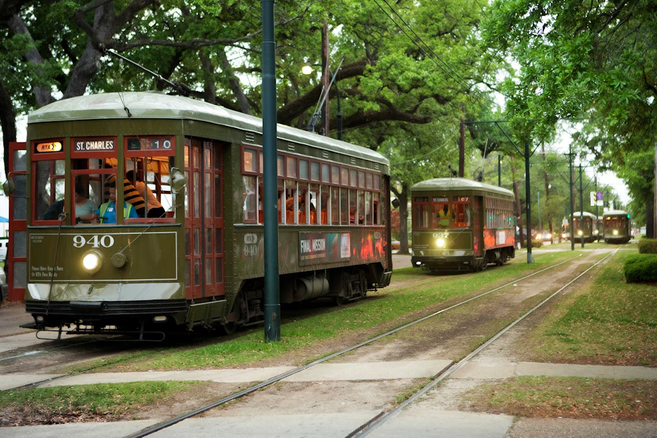 Two vintage streetcars on a green-lined track, surrounded by trees and grassy areas, with passengers visible inside.