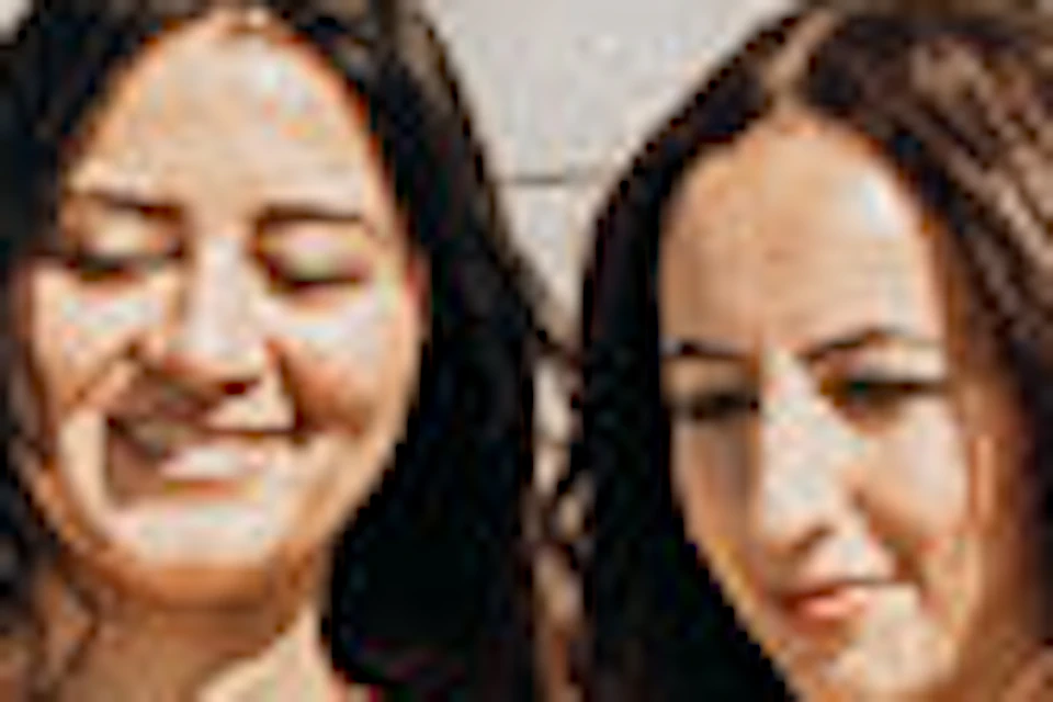 Two women pose for a photo in front of a stone monument while a person lies on the ground taking their picture with a smartphone.
