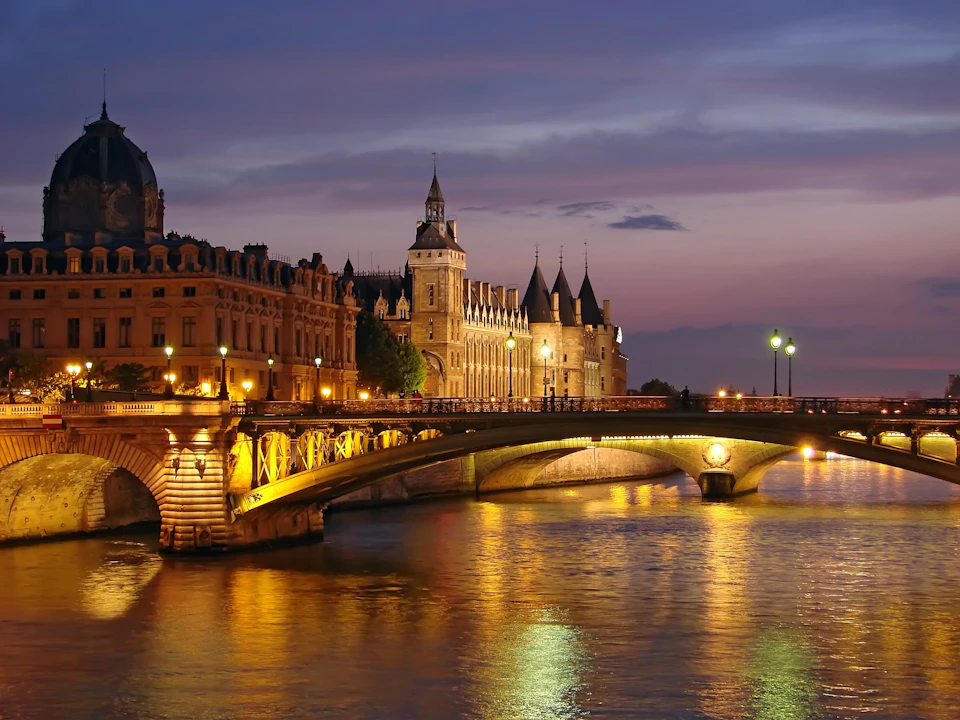 A historic bridge and lit-up buildings reflect on a river at dusk with a purple sky in the background.