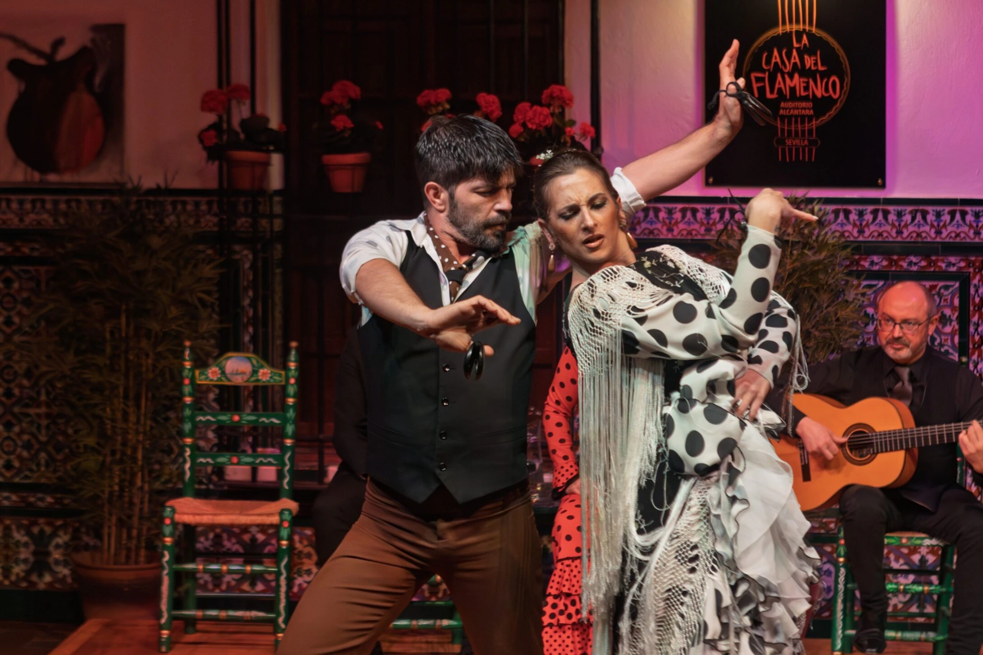 Two flamenco dancers in traditional attire perform passionately on stage, accompanied by a guitarist in the background.