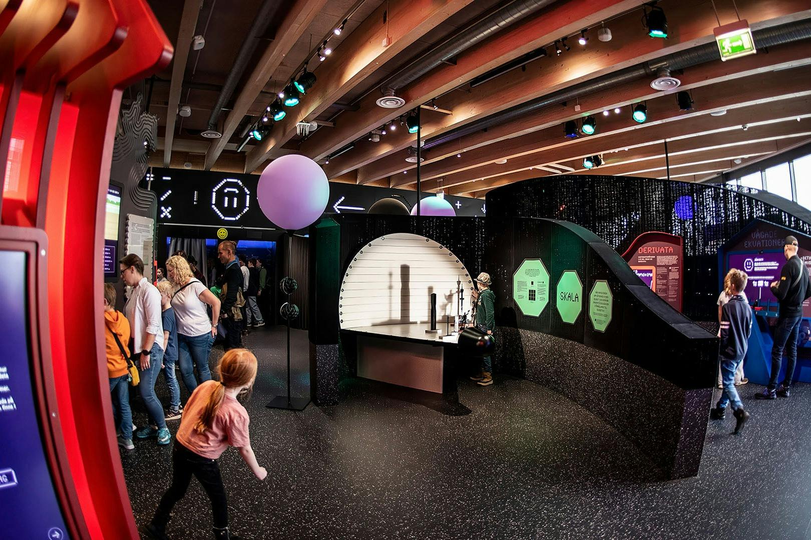 Lidé objevující interaktivní vědeckou výstavu se světly, stíny a vývěskami. V popředí kráčí dívka v růžové košili.