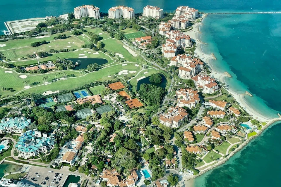 Aerial view of a coastal area with residential buildings, golf courses, tennis courts, lush greenery, and turquoise waters.