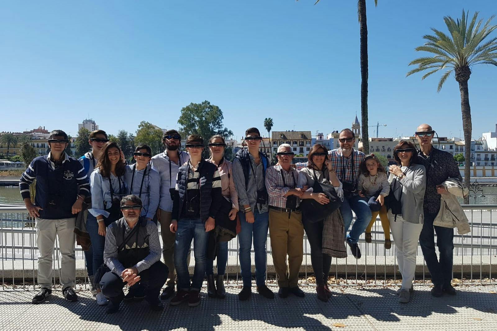 Gruppo di persone in piedi, in posa davanti a un paesaggio urbano sul lungomare con palme ed edifici sotto un cielo azzurro e limpido.