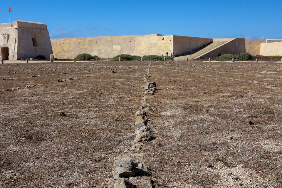 A dry, barren ground with a line of stones leading to a stone fort wall under a clear blue sky.