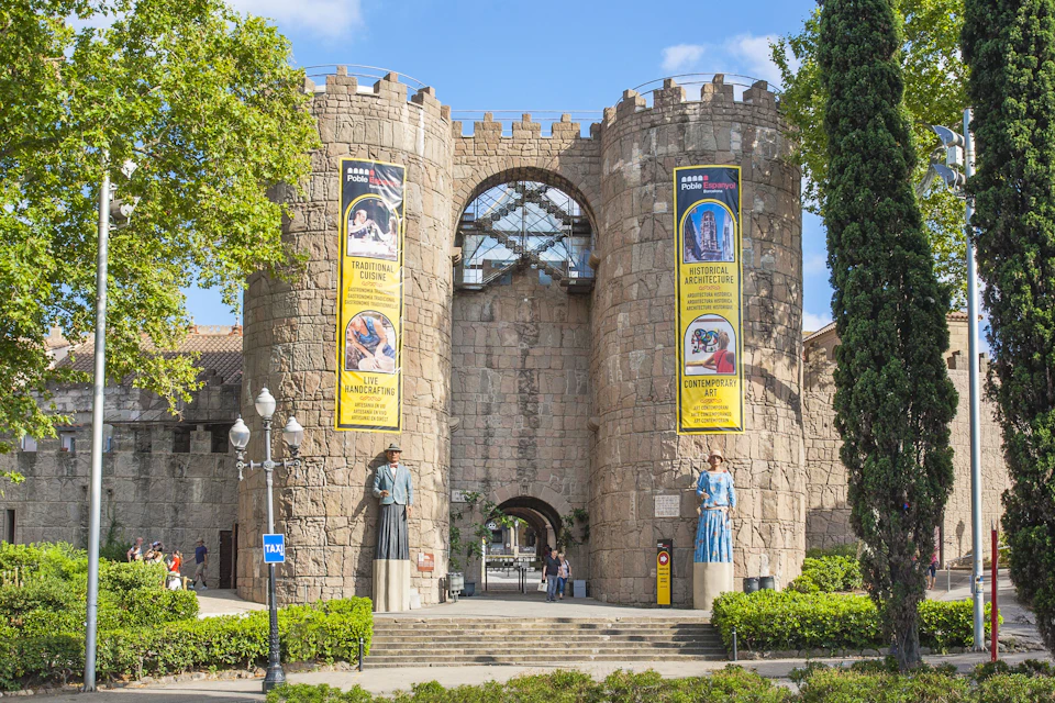 Stone entrance with two towers, colourful banners, statues, trees, and people walking through. Clear skies in the background.