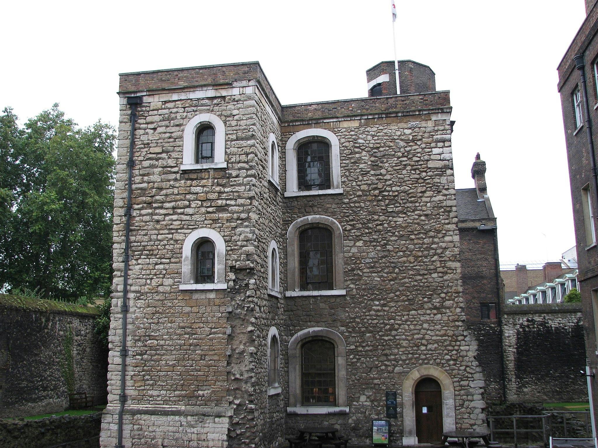 Un bâtiment en pierre avec des fenêtres cintrées et une porte en bois, entouré d'arbres et d'autres structures historiques.