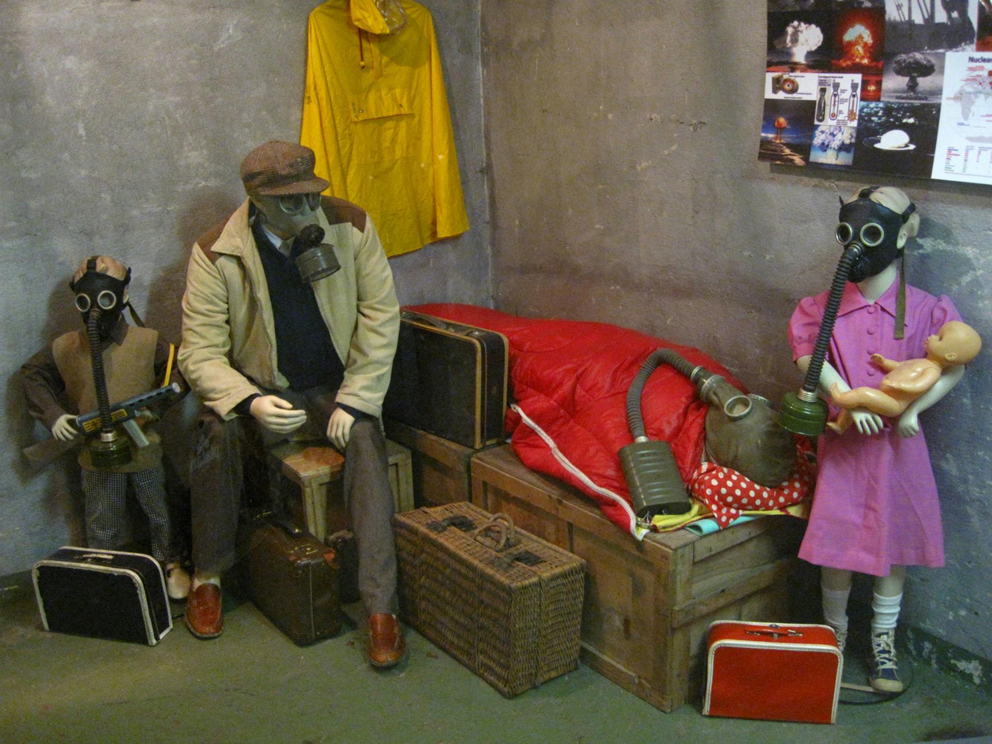 Dvě figuríny v plynových maskách sedí v místnosti s kufry a žlutou pláštěnkou zavěšenou na zdi. Na dřevěné krabici je červený spací pytel.