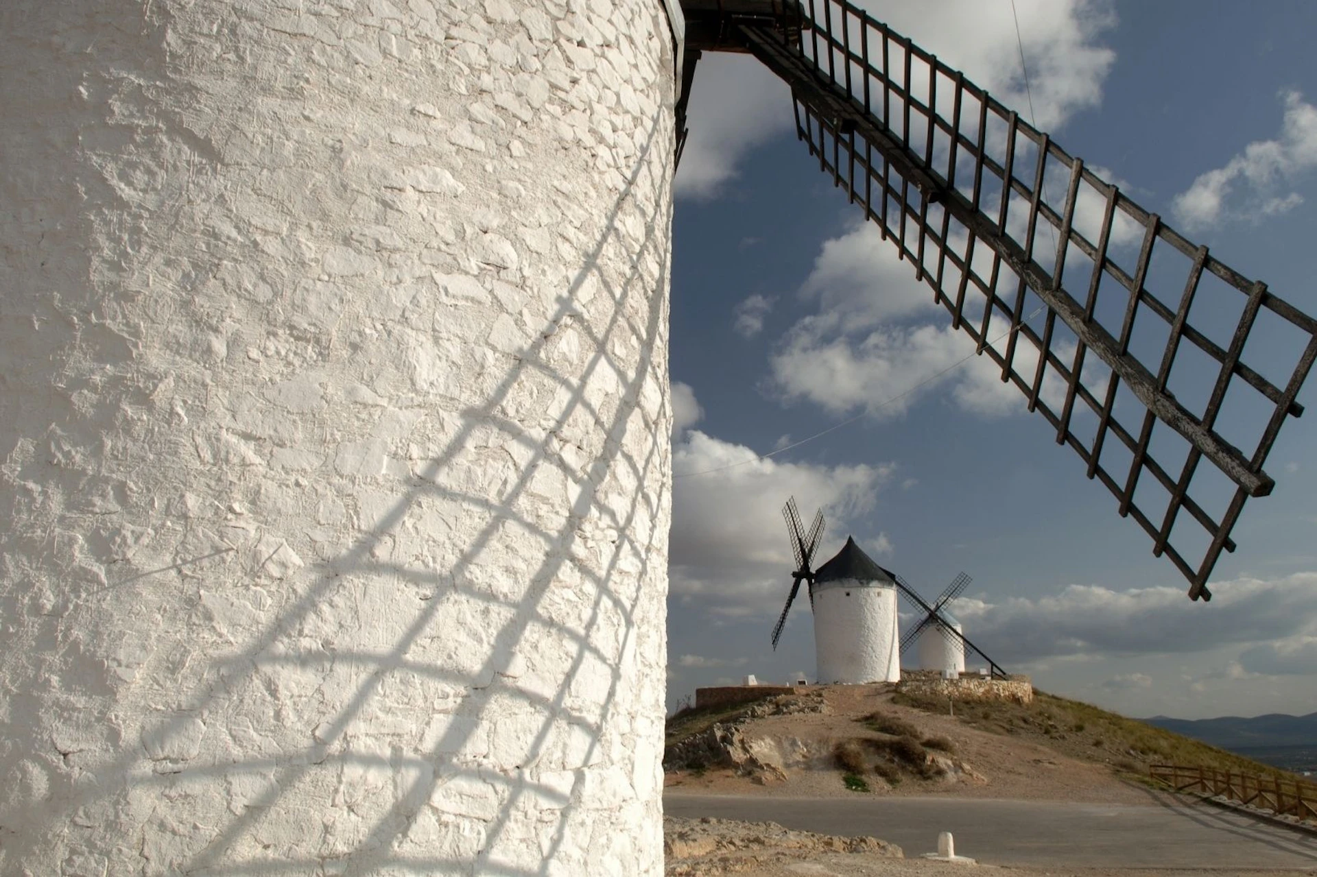 Close-up of a white windmill with its shadow on the wall, with two other windmills in the background under a cloudy sky.