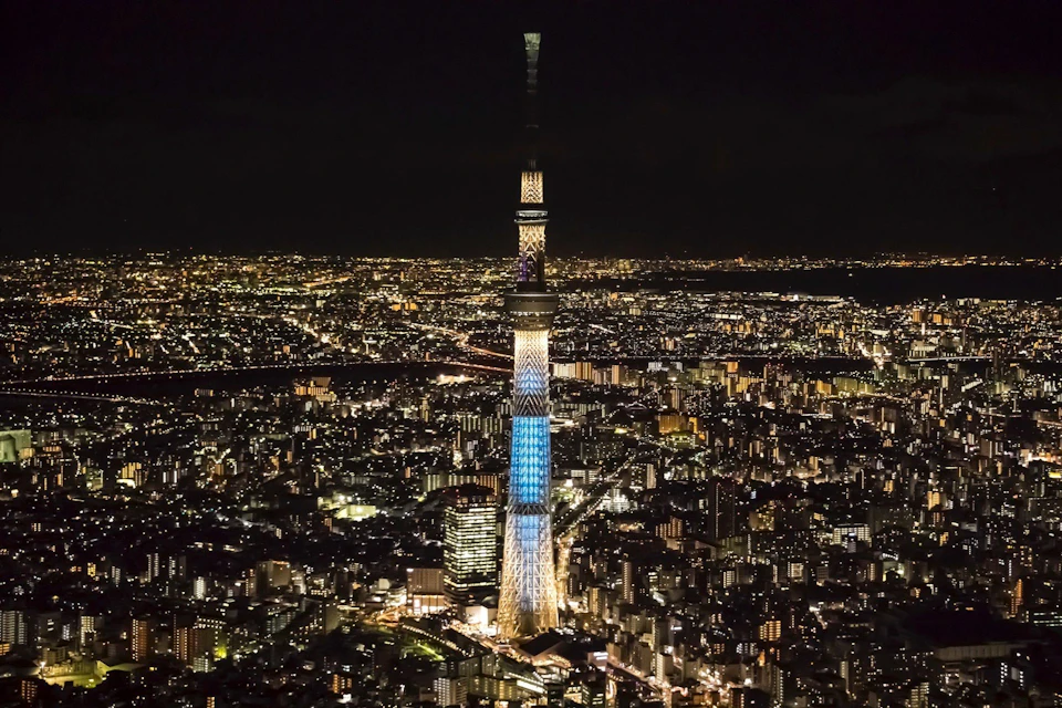 A night view of a brightly illuminated tower in a cityscape, with surrounding buildings and streets lit up.