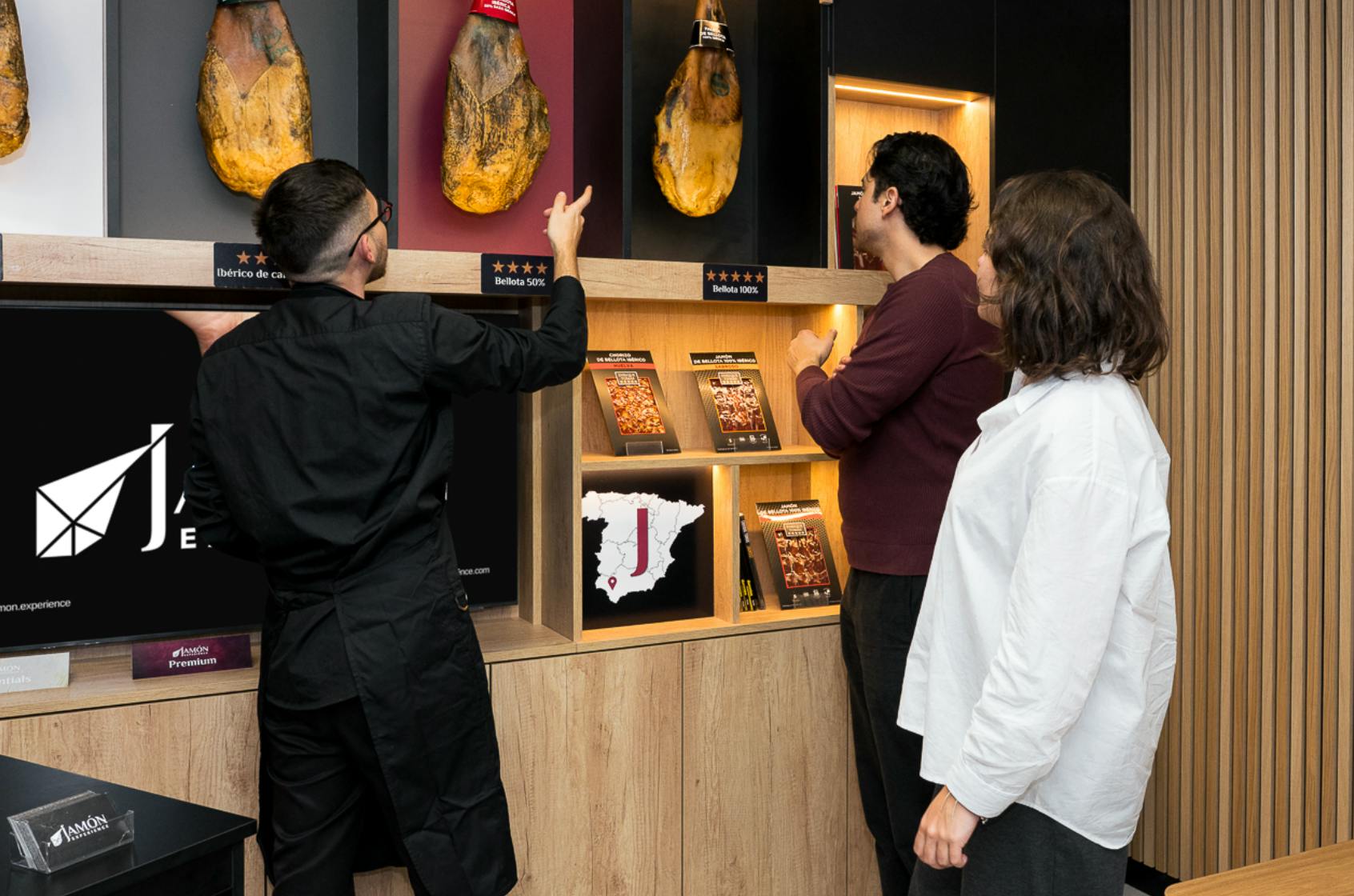 Três pessoas observam uma exposição de presunto e materiais informativos numa loja moderna.