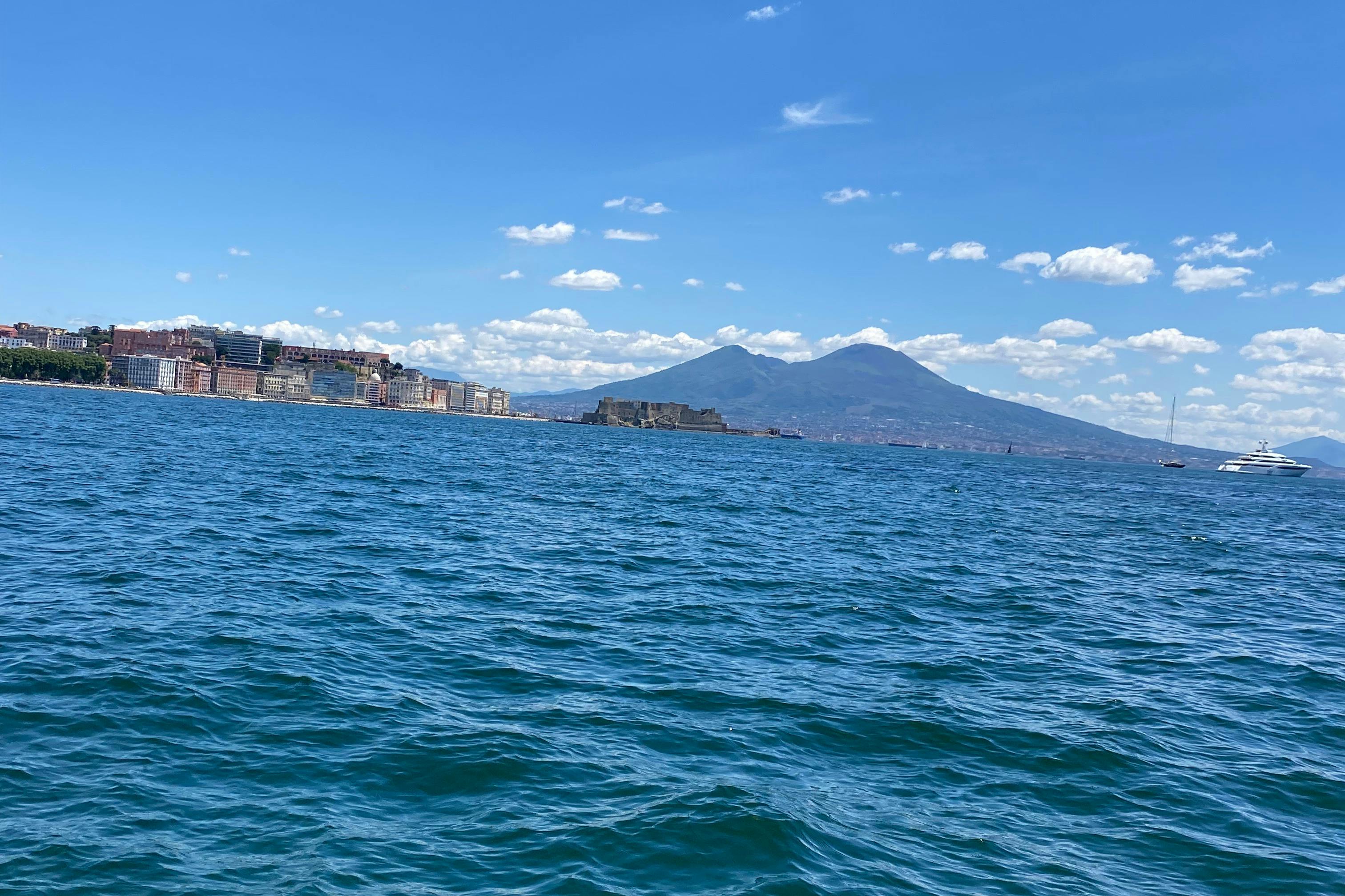 Paisaje urbano costero con edificios en una costa, un vasto mar azul y un fondo montañoso bajo un cielo despejado con nubes.