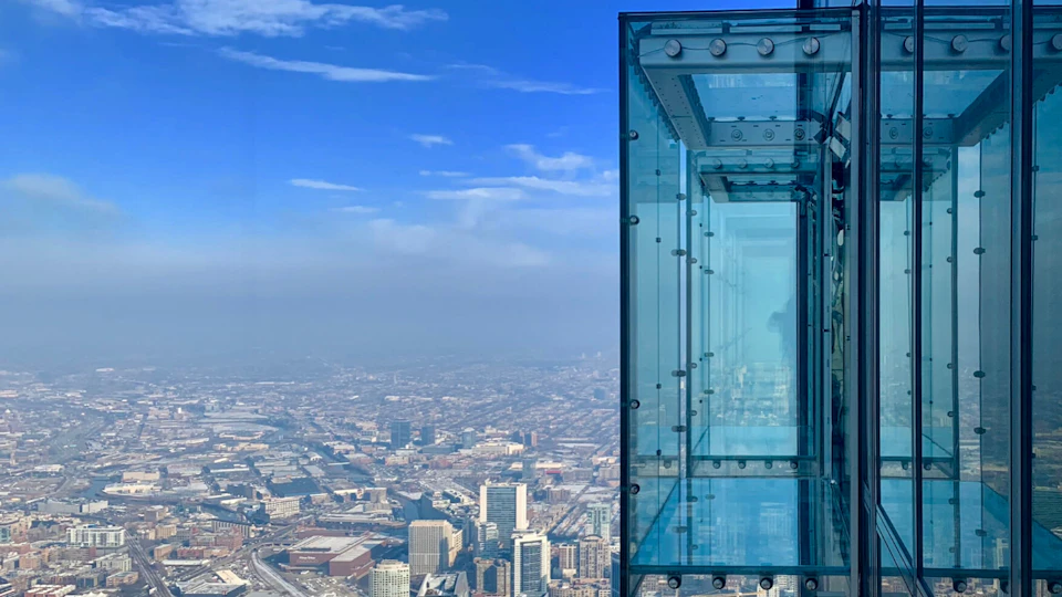 A glass observation deck juts out from a high-rise building, overlooking a cityscape with clear skies.