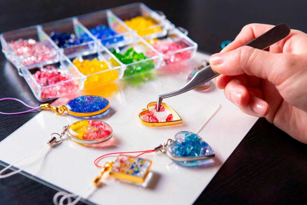A hand uses tweezers to place colorful resin pieces into a heart-shaped frame, with other jewelry and materials nearby.