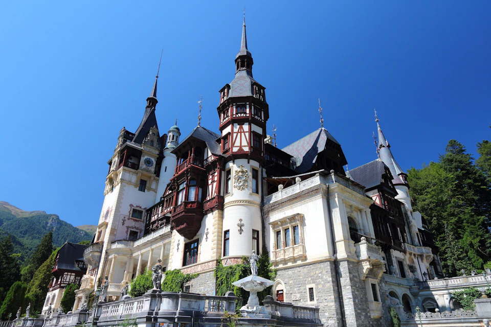 A grand castle with pointed towers, intricate details, and a stone base under a clear blue sky. A fountain is visible in front.