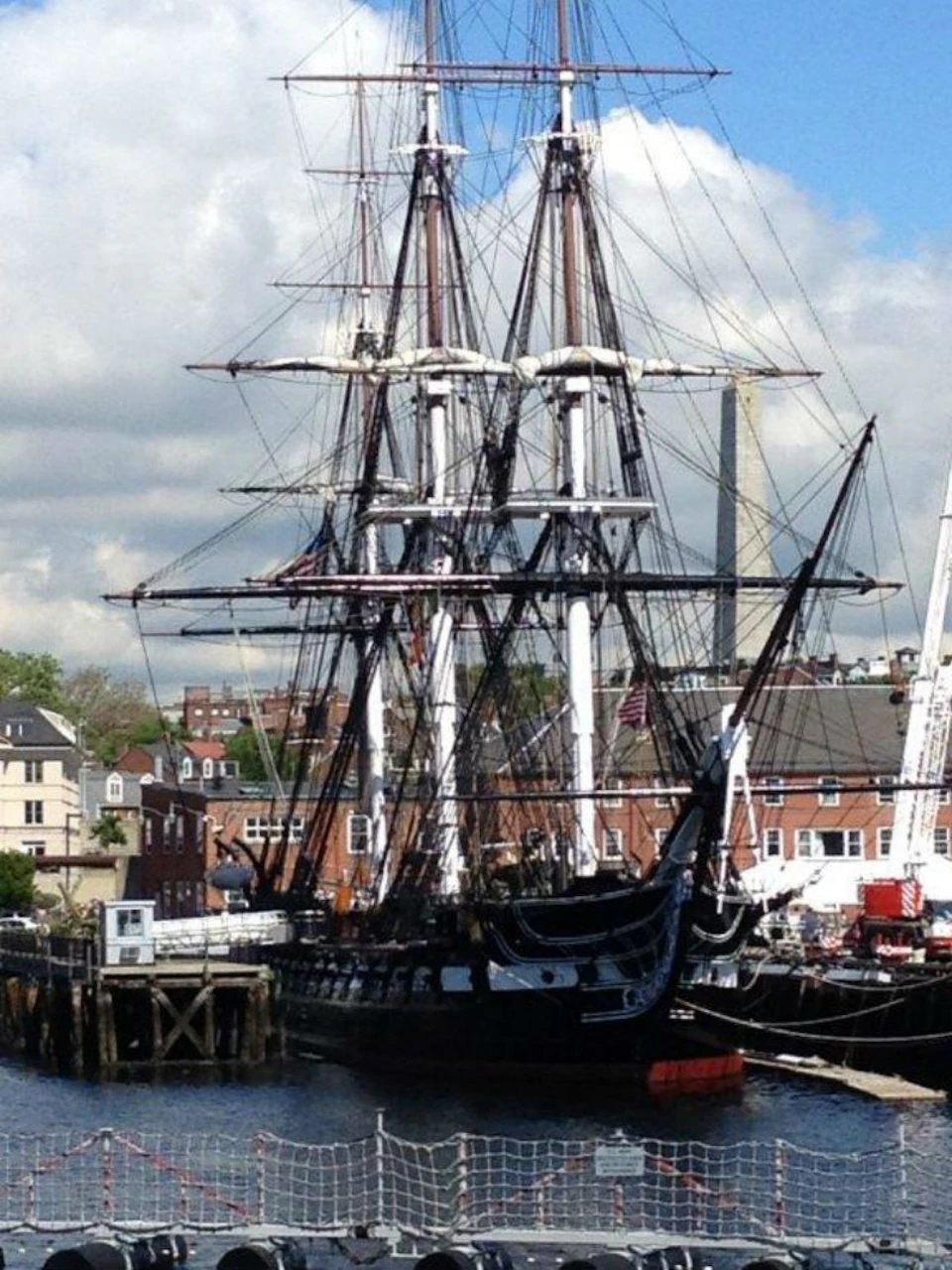 Historic sailing ship docked at a pier with buildings and a large obelisk visible in the background under a partly cloudy sky.