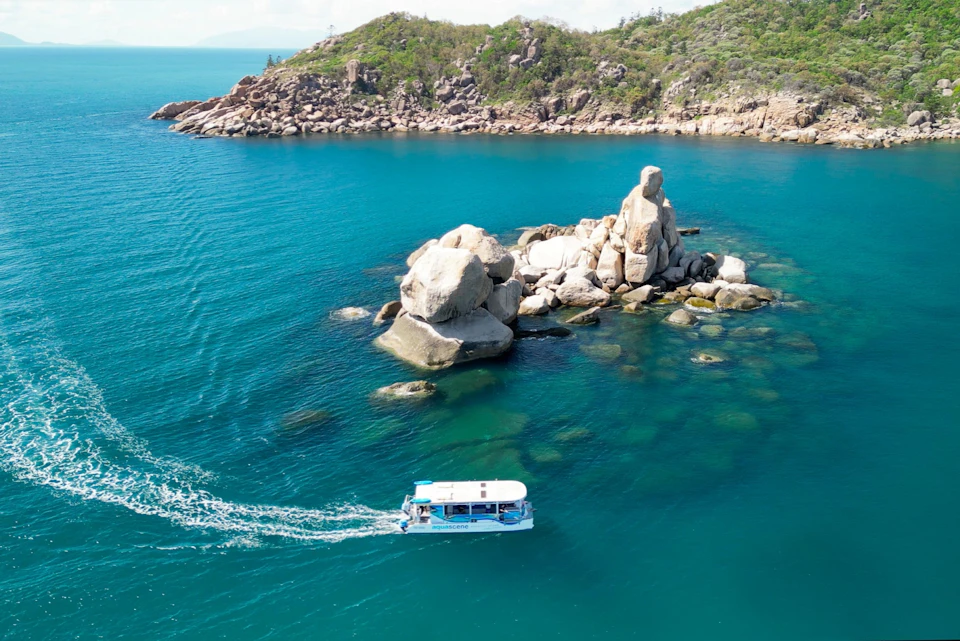 A small boat sails near rocky islets surrounded by blue water, with a green, rocky shoreline in the background.